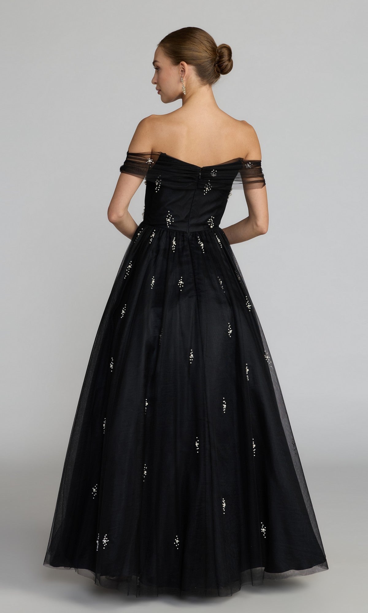 A woman stands with her back to the camera against a plain background, wearing the Mac Duggal 13227 long formal dress—an off-the-shoulder black gown featuring a full skirt and embellished details.